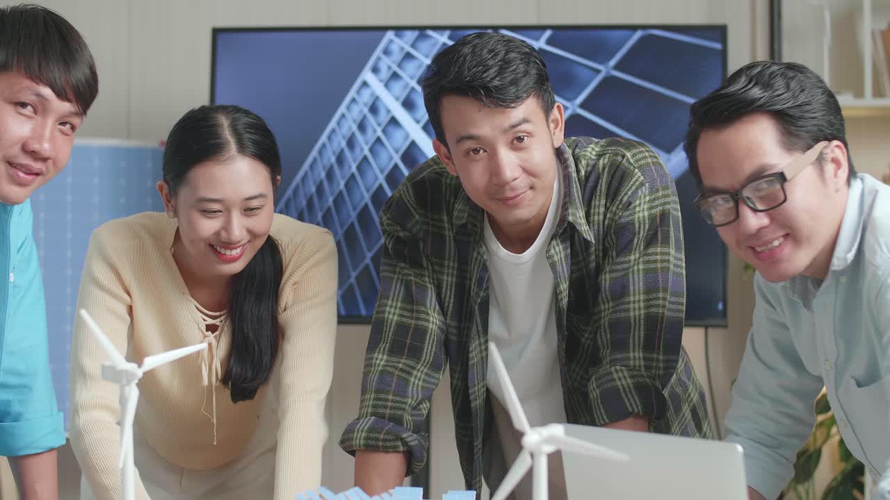 사무실에서 태양광 패널을 가진 작은 집 모델에 대해 일하는 동안 카메라에 미소 짓는 아시아 엔지니어 그룹의 클로즈업