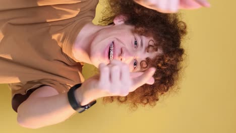 cheerful young man shows rock hand gesture with playful expression indoors a young man with a mix of african and caucasian features smiles widely sticking out his tongue while making a rock hand gesture the vibrant yellow background adds energy to the moment