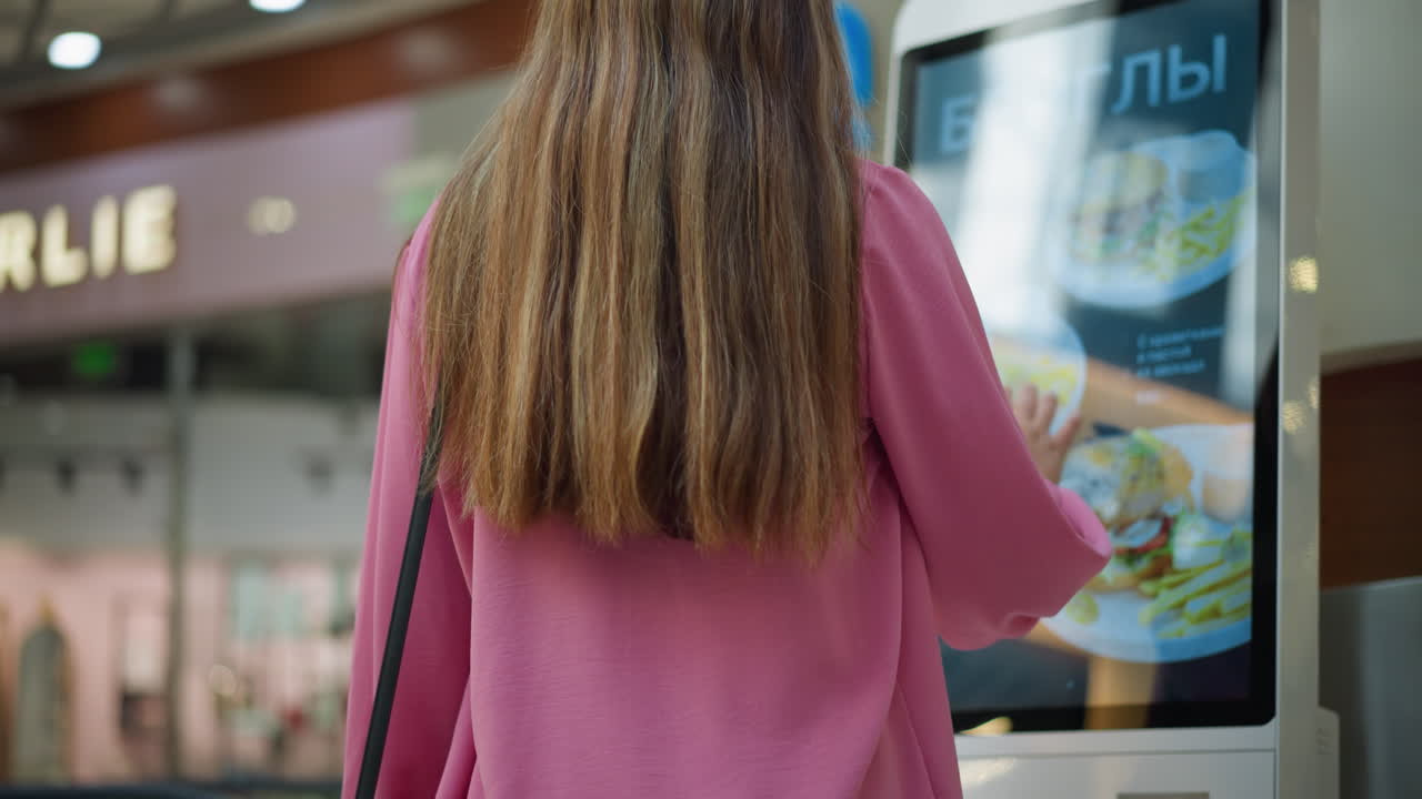 vista trasera de una dama en vestido rosa con un bolso negro interactuando con una máquina de pedidos de alimentos de pantalla táctil en un centro comercial bien iluminado, se puede ver un ligero borrón de personas y elementos de fondo