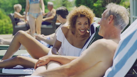 retired senior couple on loungers relaxing by swimming pool on summer vacation