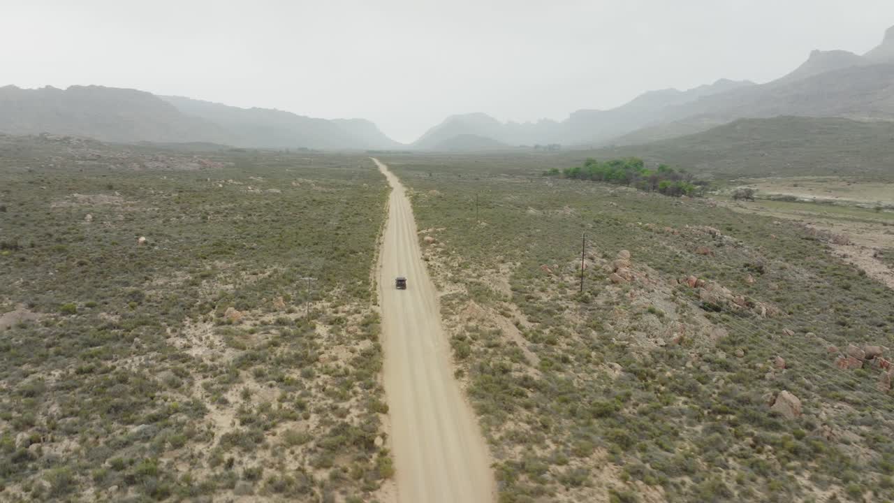 드론은 도로 근처에서 높이 날아와 새의 눈으로 검은색 오프로드 차량을 따라갑니다. - 남아프리카 공화국의 세더버그 야생 지역의 사막 풍경 - 지평선에서 산맥을 볼 수 있습니다.