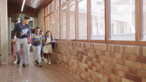 Happy-diverse-male-teacher-and-children-with-books-in-elementary-school-corridor,-slow-motion