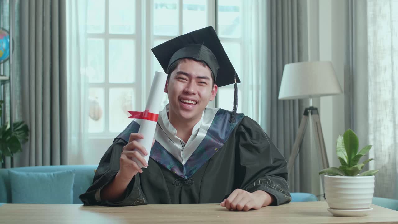 オンラインビデオコールで大学卒業証書を披露する興奮したアジア人男性卒業式のドレスとキャップを着てリビングに座っている男性卒業生