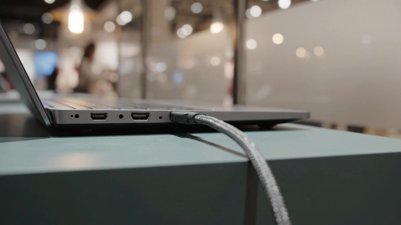 Primo piano dettagliato della vista laterale di un laptop che mostra le porte e una connessione via cavo USB-C su un moderno tavolo espositivo in uno spazio espositivo luminoso.