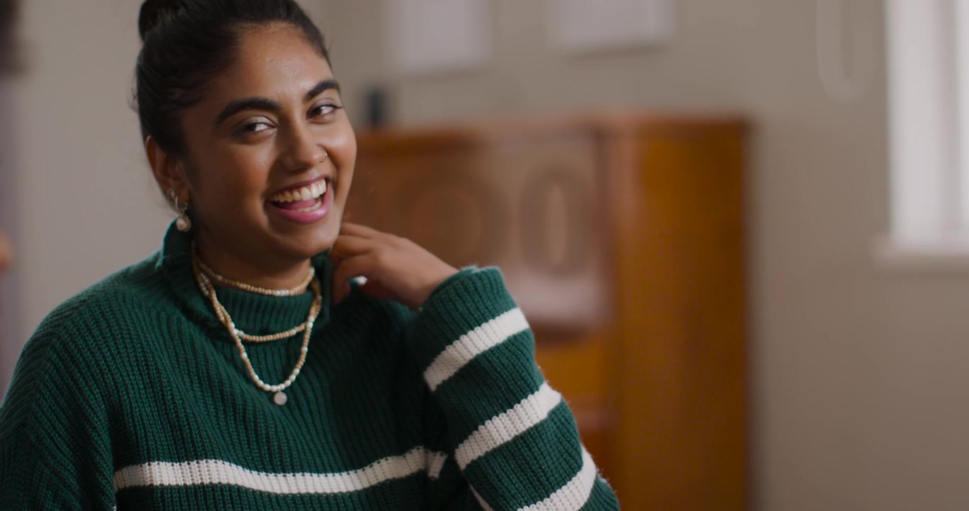 Indian woman in green knit laughing touching necklace while friend talking at table, copy space