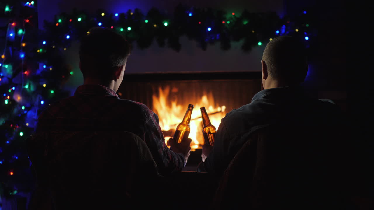 두 남자는 벽난로와 크리스마스 장식 옆에서 맥주 한 병을 손에 들고 휴식을 취합니다.