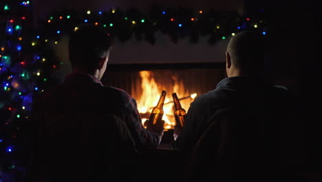 two men rest with bottles of beer in hand by the fireplace and christmas decorations