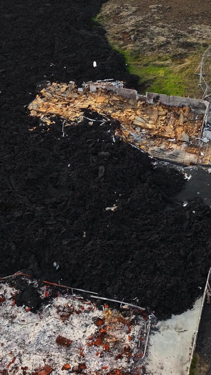 Vertical drone view of hardened lava engulfing destroyed buildings in Grindavik, Iceland, with black volcanic rock surrounding exposed timber and masonry debris.