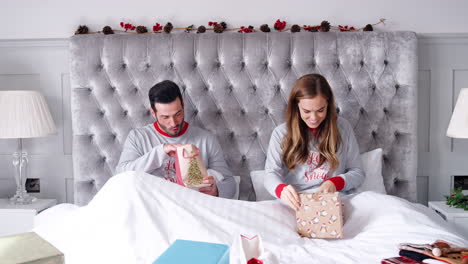 woman opening gift of necklace in bed at home as couple exchange presents on christmas day