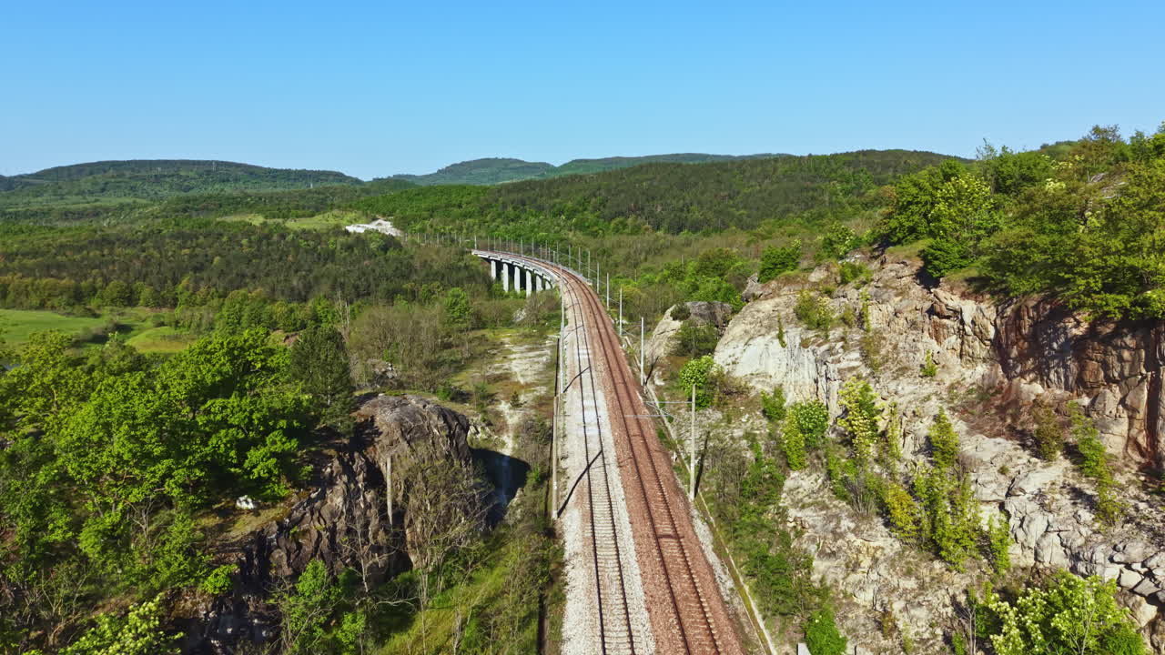 I binari del treno si estendono su un ponte che attraversa una valle con colline verdi e alberi, sotto un cielo azzurro e limpido, di giorno