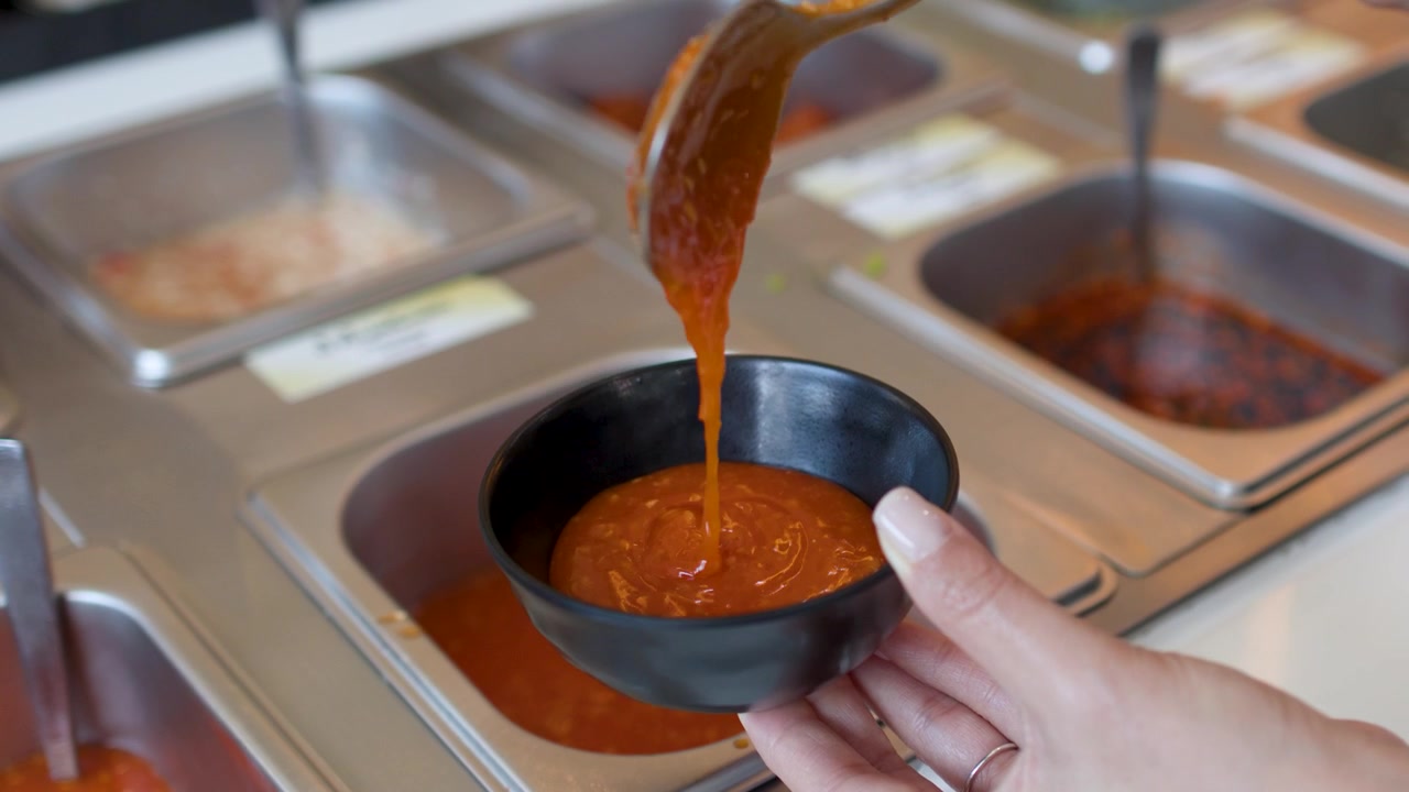 Nahaufnahme von Händen, die roten Chili-Dip von einer Buffetstation in eine Schüssel schöpfen.