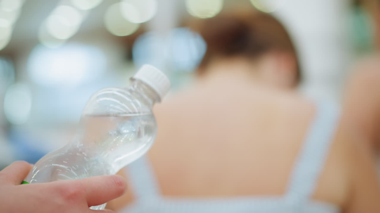 una mujer sostiene agua en botella fría en un centro comercial detrás de otra mujer, sacudiendo gradualmente la botella, el fondo presenta iluminación borrosa, y la botella es clara y fría con condensación visible