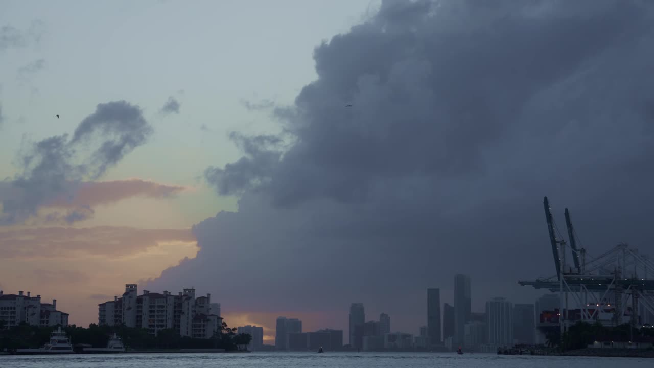 A dramatic wall of clouds approaches the Miami skyline during sunset, with cranes silhouetted against the fading light.