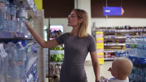 Attractive-woman-in-glasses-is-choosing-a-bottle-of-water-in-beverages-department-in-the-supermarket,-while-her-little-baby-is