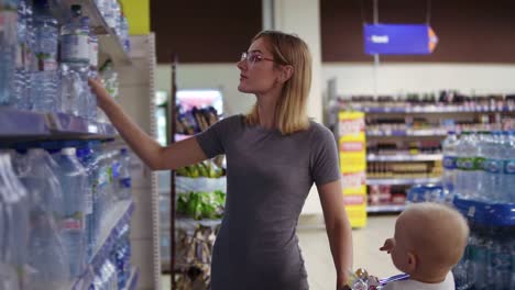 mujer atractiva con gafas está eligiendo una botella de agua en el departamento de bebidas en el supermercado, mientras que su pequeño bebé es
