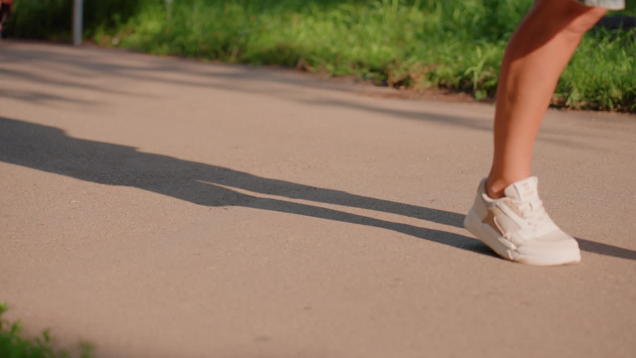 Sendero iluminado por el sol en un parque con zapatillas y rueda de bicicleta pasando, peatones deteniéndose en pavimento cálido cerca del borde verde; primeros planos de suelas de zapatos, sombras extendiéndose por el asfalto, pasos lentos