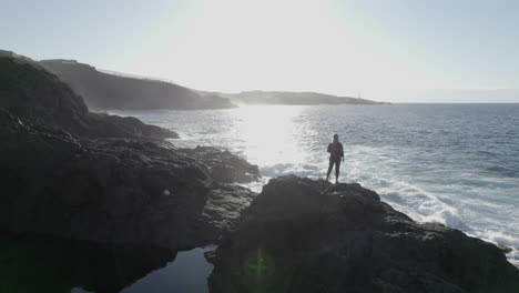 aerial shot of a young woman in orbit that is on a rock near the sea and waves