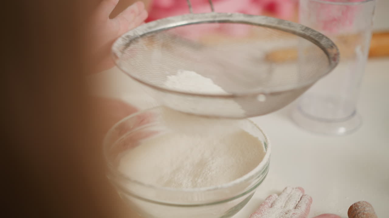 close up of flour being sifted through fine metal sieve into transparent glass bowl on white kitchen table, powder gently falling while child hand rests on surface
