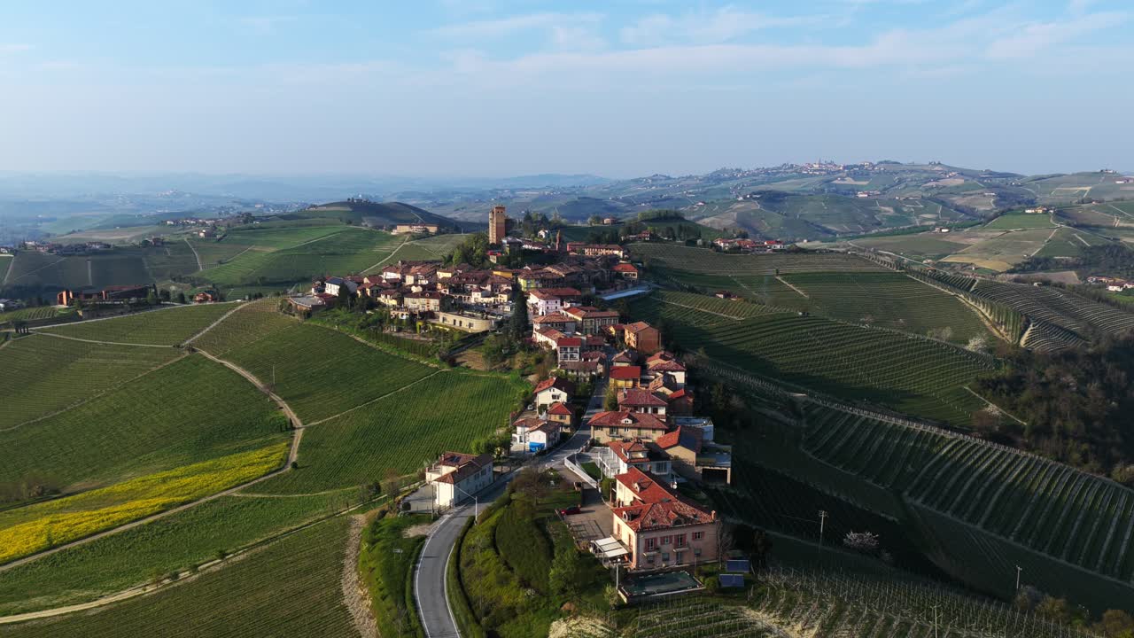 Orbiting aerial shot of Serralunga d'Alba, capturing its medieval castle and clustered village surrounded by rolling vineyards, with sweeping views over the scenic Langhe hills under a clear sky