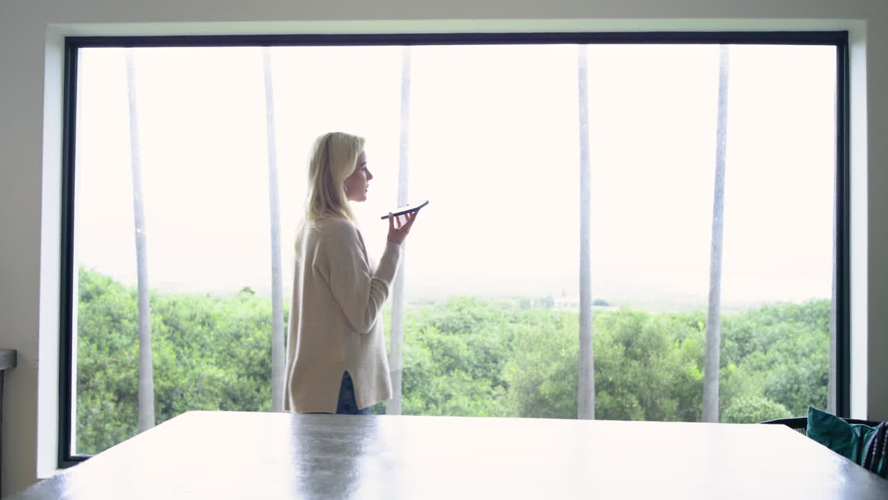 White woman making smartphone call while moving along window ledge in room delivering final remark