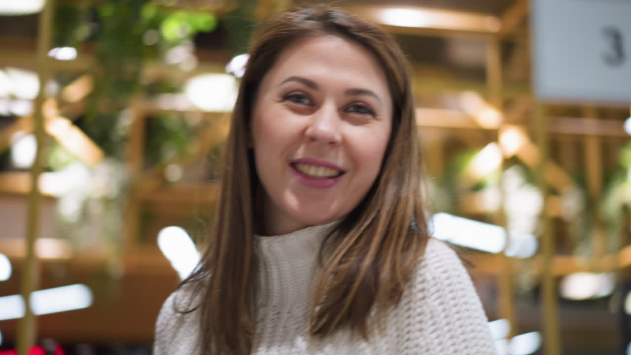 primer plano de una joven mujer caucásica en un suéter blanco, sonriendo sutilmente a la cámara frente a una tabla de salida del aeropuerto borrosa