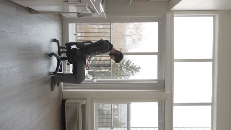 vertical view, young male, gamer or student, working on laptop in chair in room on daylight with headphones on head, static shot