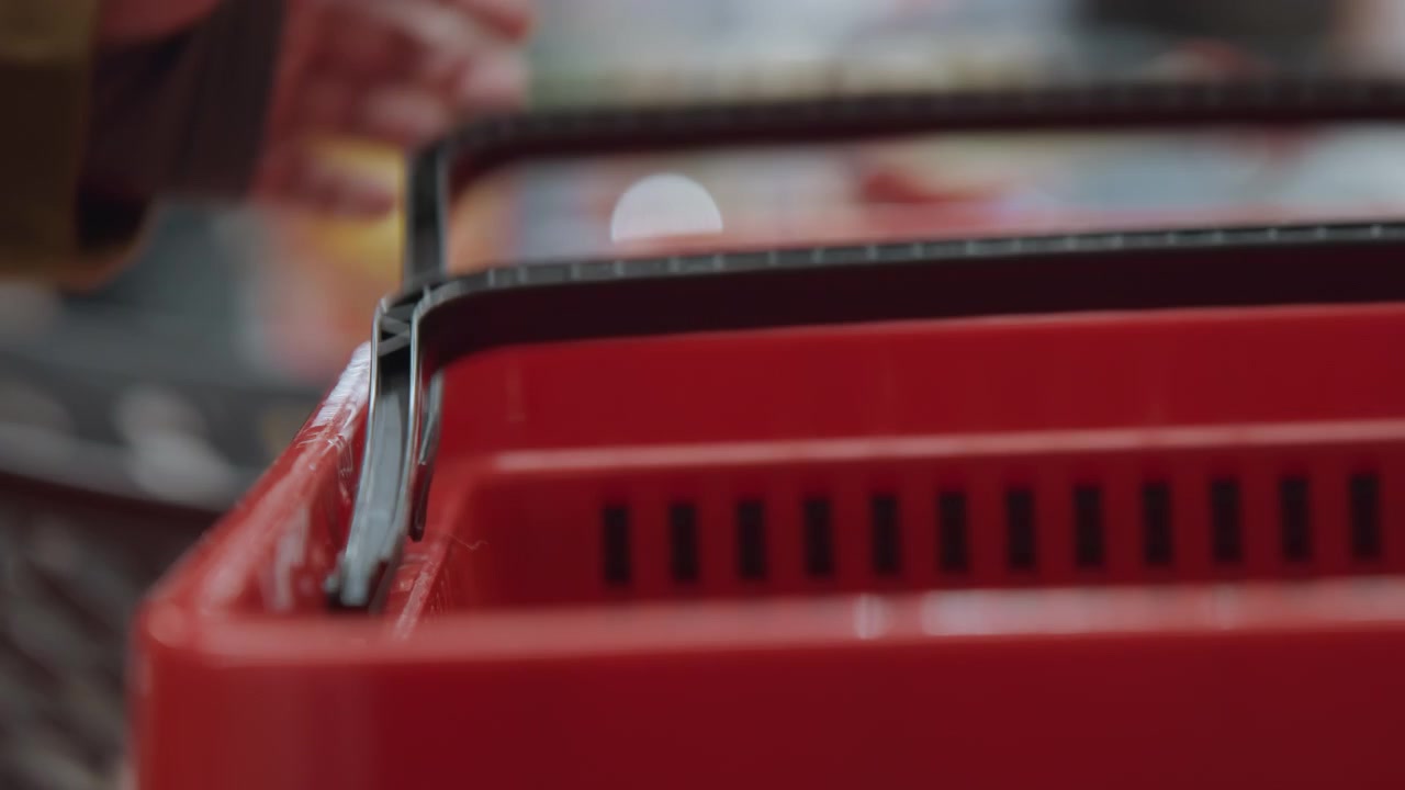 Red shopping basket handles in supermarket, closeup shallow depth of field showing worn black handle and plastic grid, blurred shoppers in background, shopper lifts basket by handle, muted fluorescent