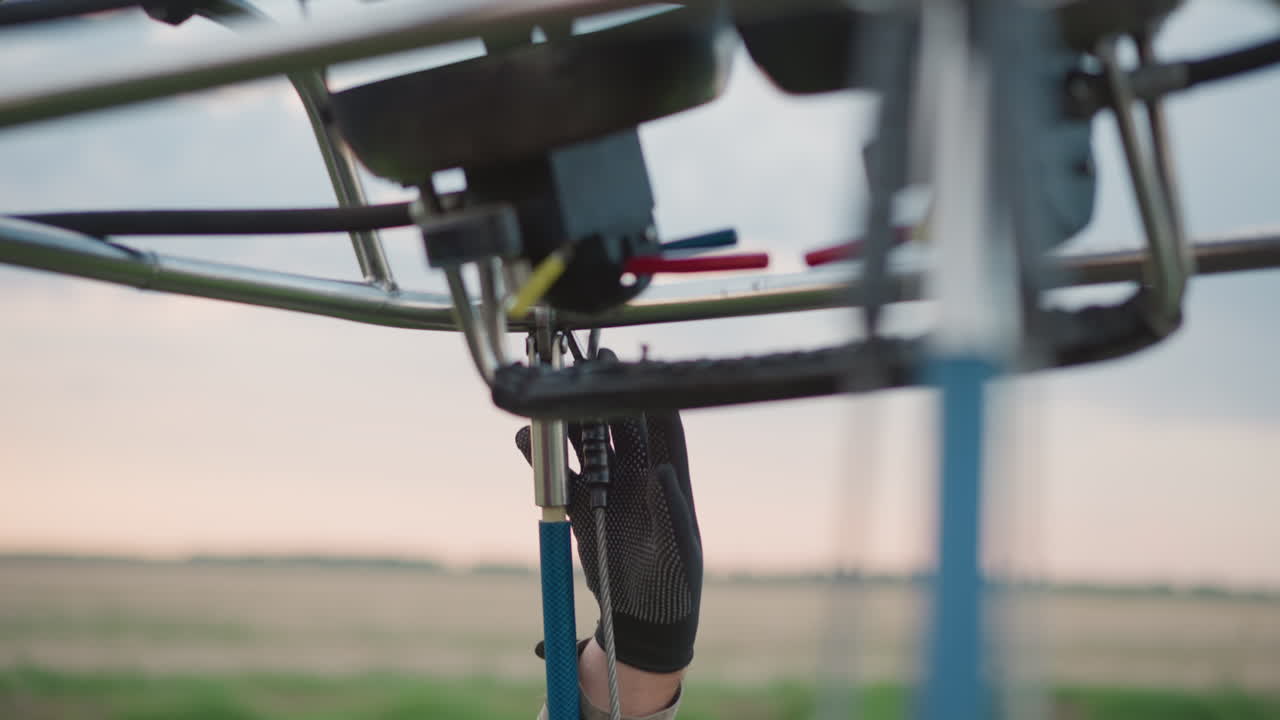 gloved hands securing bolt hook on burner frame of hot air balloon during preflight setup under cloudy sky with close up of hose wires and metal basket edge visible regulator gauge pressure detail
