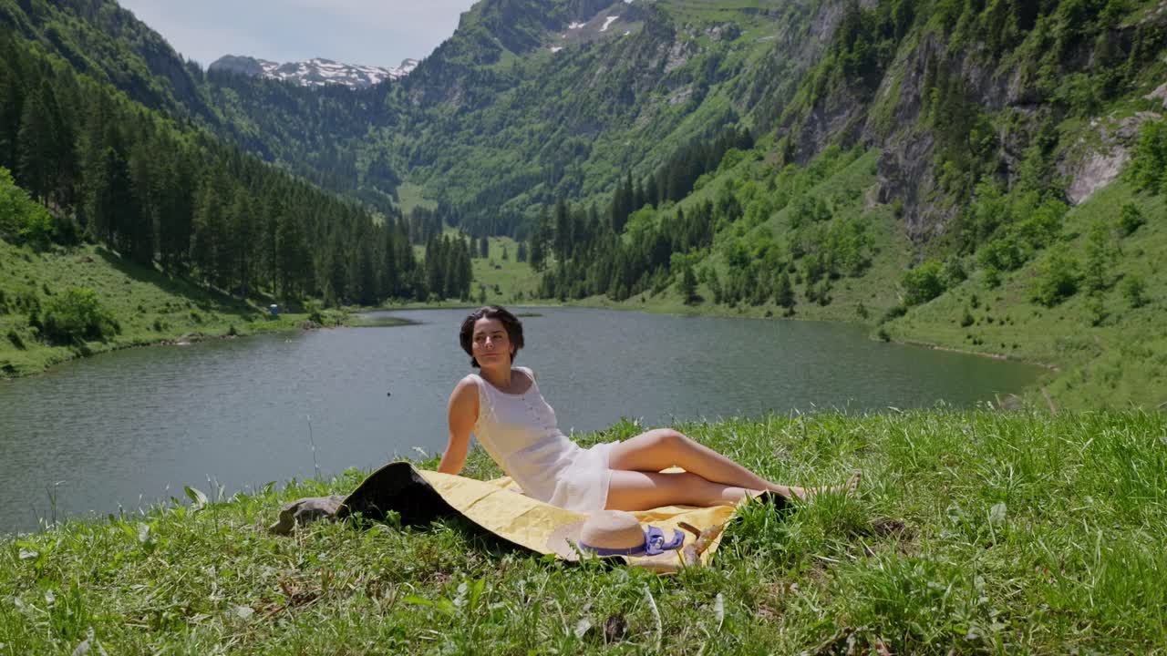 Pretty Woman on picnic blanket enjoying sun in Swiss mountains. Beautiful tranquil lake in background. Dolly portrait shot. Summer season between hills.