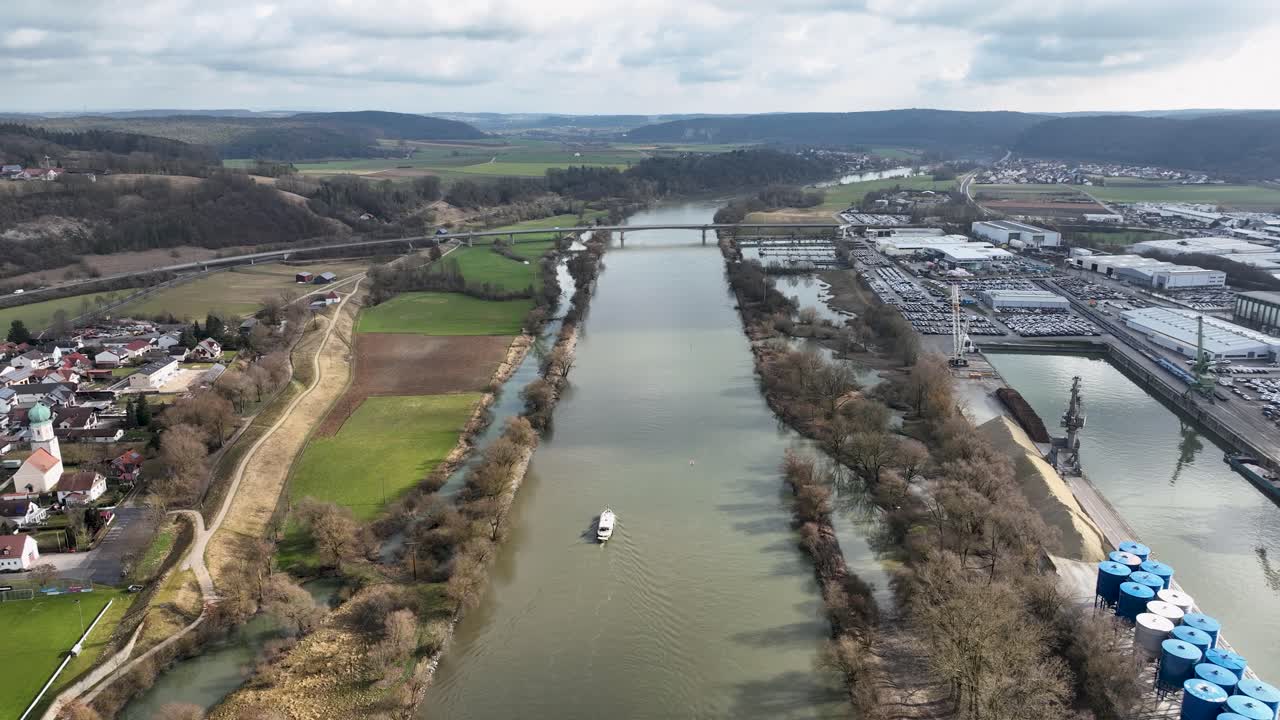 A drone perspective of a barge traveling on the canal in Kelheim, passing by a busy industrial harbor with parked vehicles and cranes