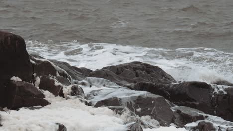 slow motion waves and sea foam gather around well worn black rocks on a stormy and unsettled day