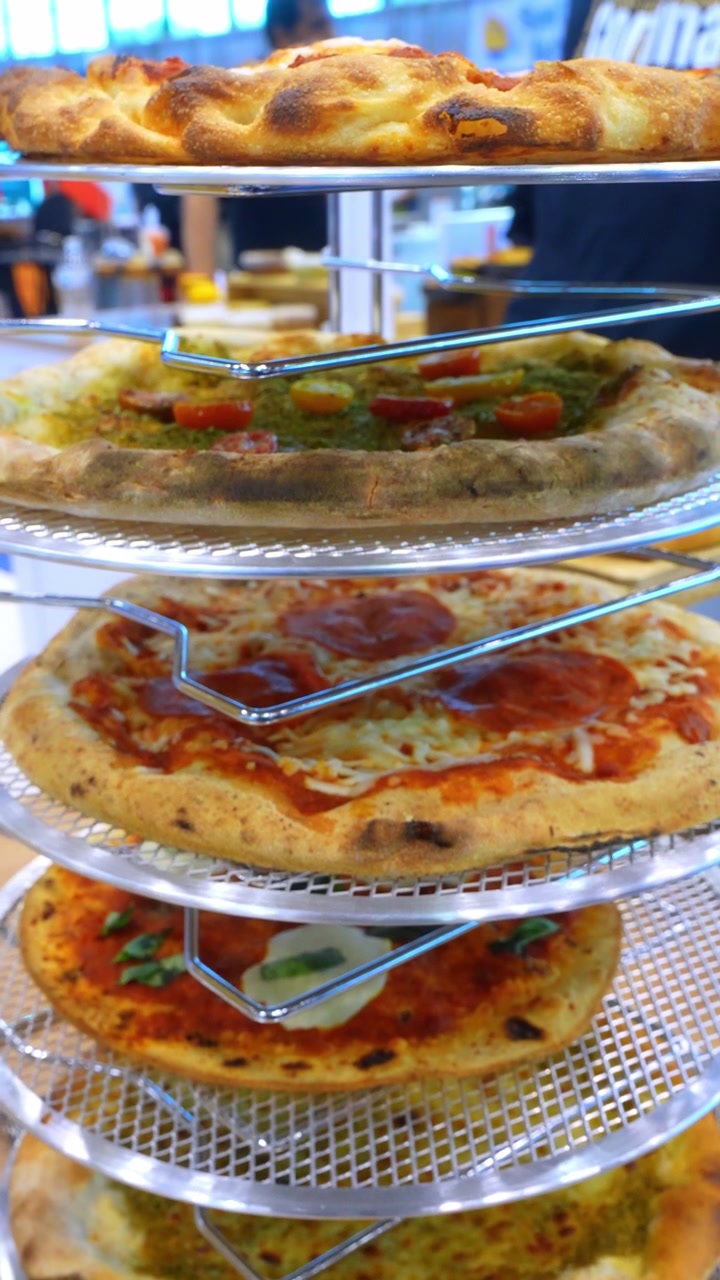Pizze assortite esposte su rastrelliere metalliche impilate. Una vista verticale ravvicinata mostra varietà di pepperoni, pomodoro, basilico e crosta ripiegata all'interno di una mostra alimentare