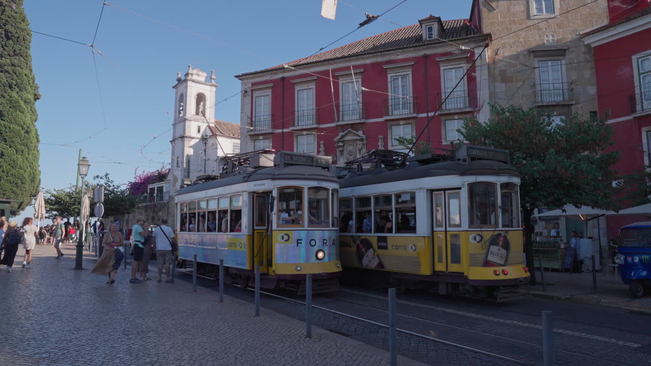 Two electric trolleys filled with people pass each other on the cobblestone streets of Lisbon, Portugal, as onlookers admire the historic surroundings and vibrant city.