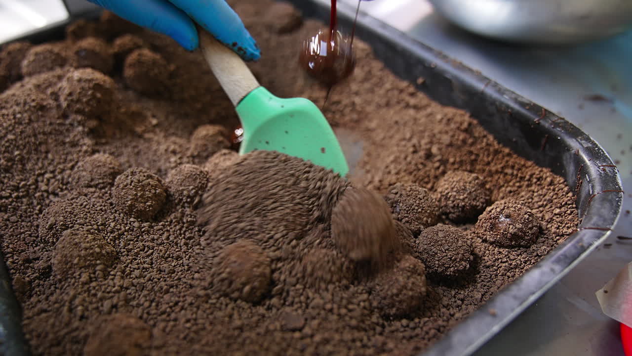 Candy balls being mixed with cocoa sprinkling. Spatula turns the sweets in a tray to create the coating. Close up.