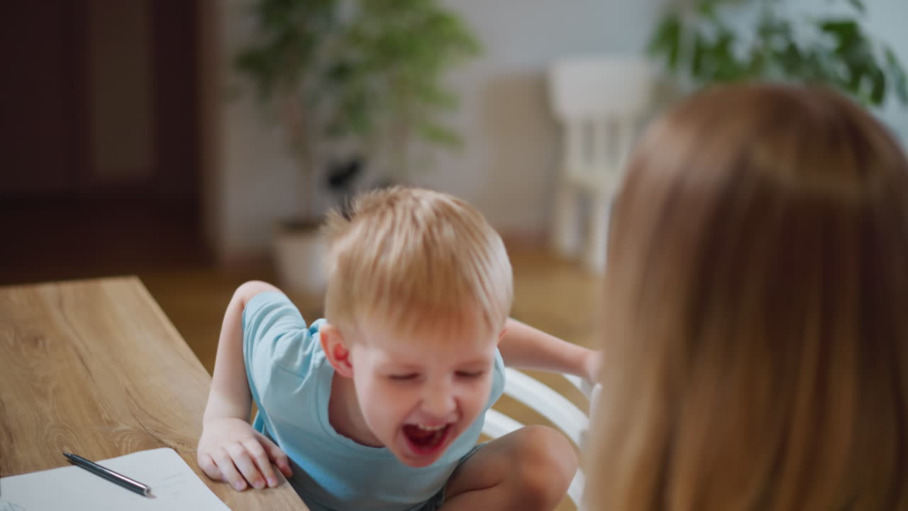 Smiling child in blue shirt leaning toward woman while sitting at table with notebook and pens, showing happiness, family connection, communication, playful interaction, bonding, learning, and joyful childhood