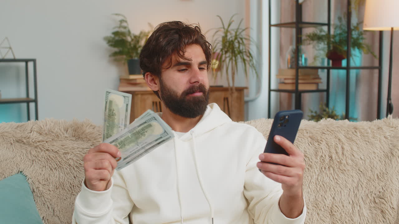 Giovane uomo indiano ricco e di successo che tiene in mano una banconota da un dollaro e uno smartphone, seduto sul divano di casa.