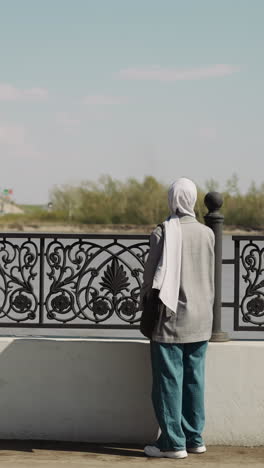 muslin woman with long hijab stands by fence on waterfront backside view. lady in jeans and jacket in town. urban architecture and islamic traditions