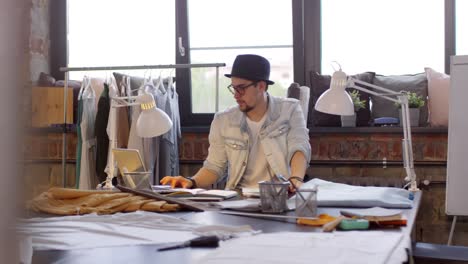 Young-Male-Fashion-Designer-Sitting-At-Desk,-Using-Laptop-And-Drawing-Sketch-Of-New-Dress-In-Modern-Sewing-Studio