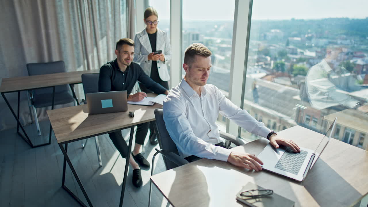 Business team in the sunny office working on computers. Lady in white jacket standing near the desk. Panoramic windows with cityscape in blur at backdrop.
