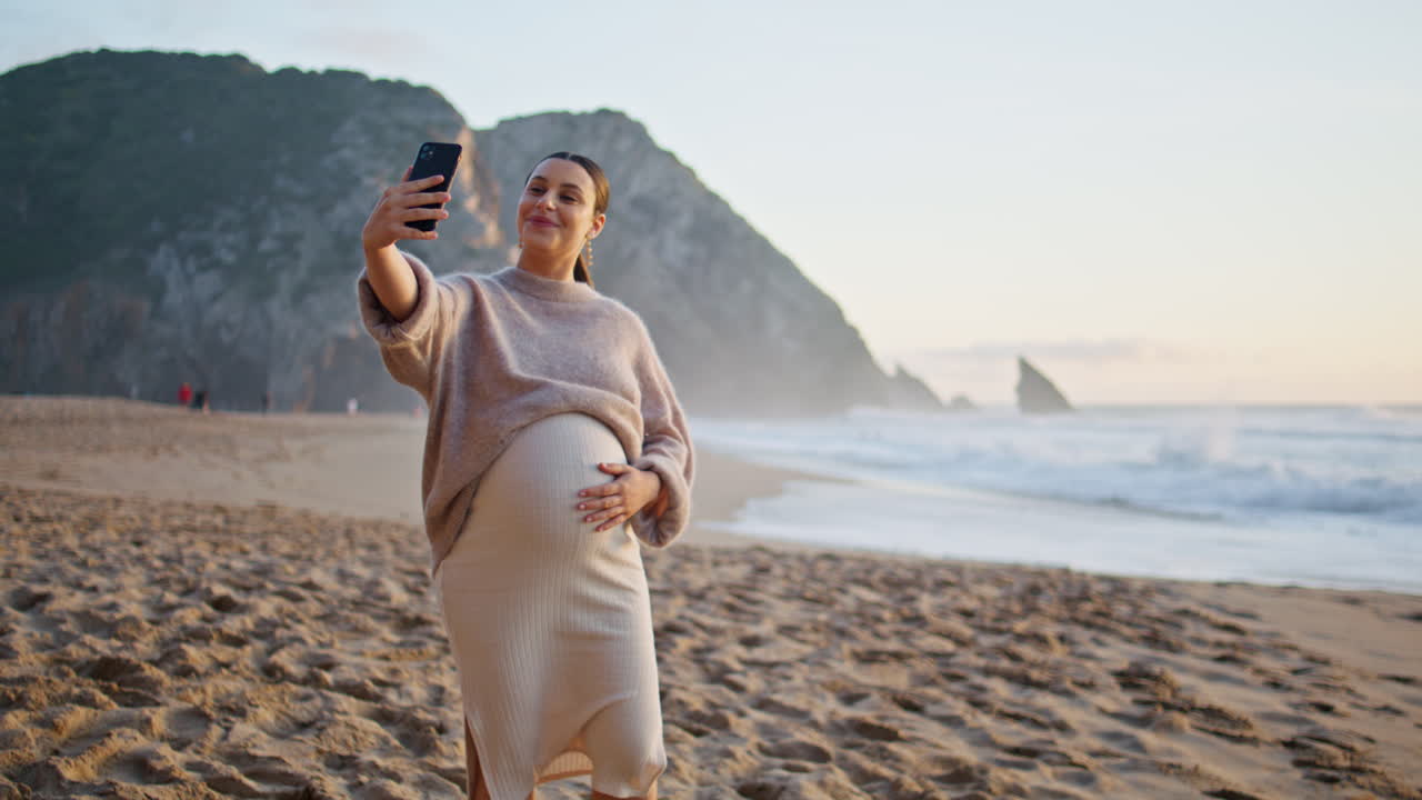 임신 중인 여성이 해변에서 휴대 전화로 비디오 통화를합니다. 행복한 임신 여성 인사
