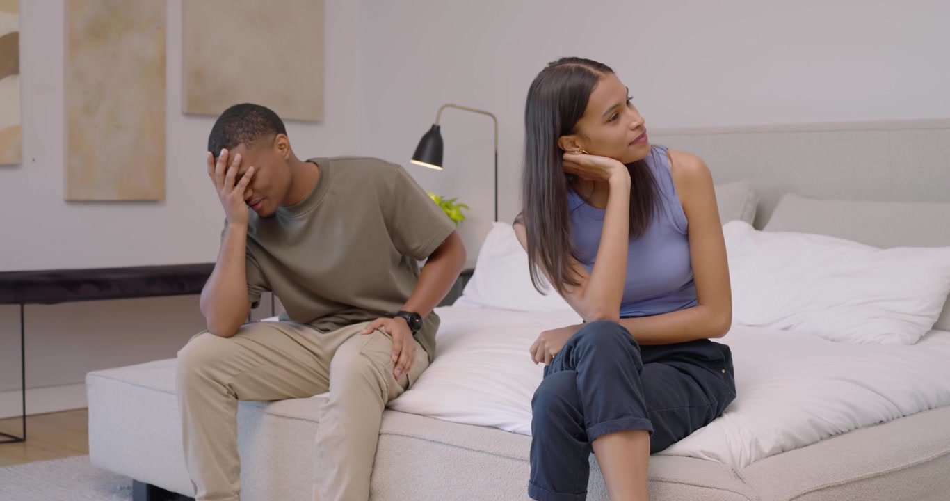 Coppia afroamericana che parla sul letto mentre l'uomo entra avvicinandosi alla donna, gesticolando e spiegando.