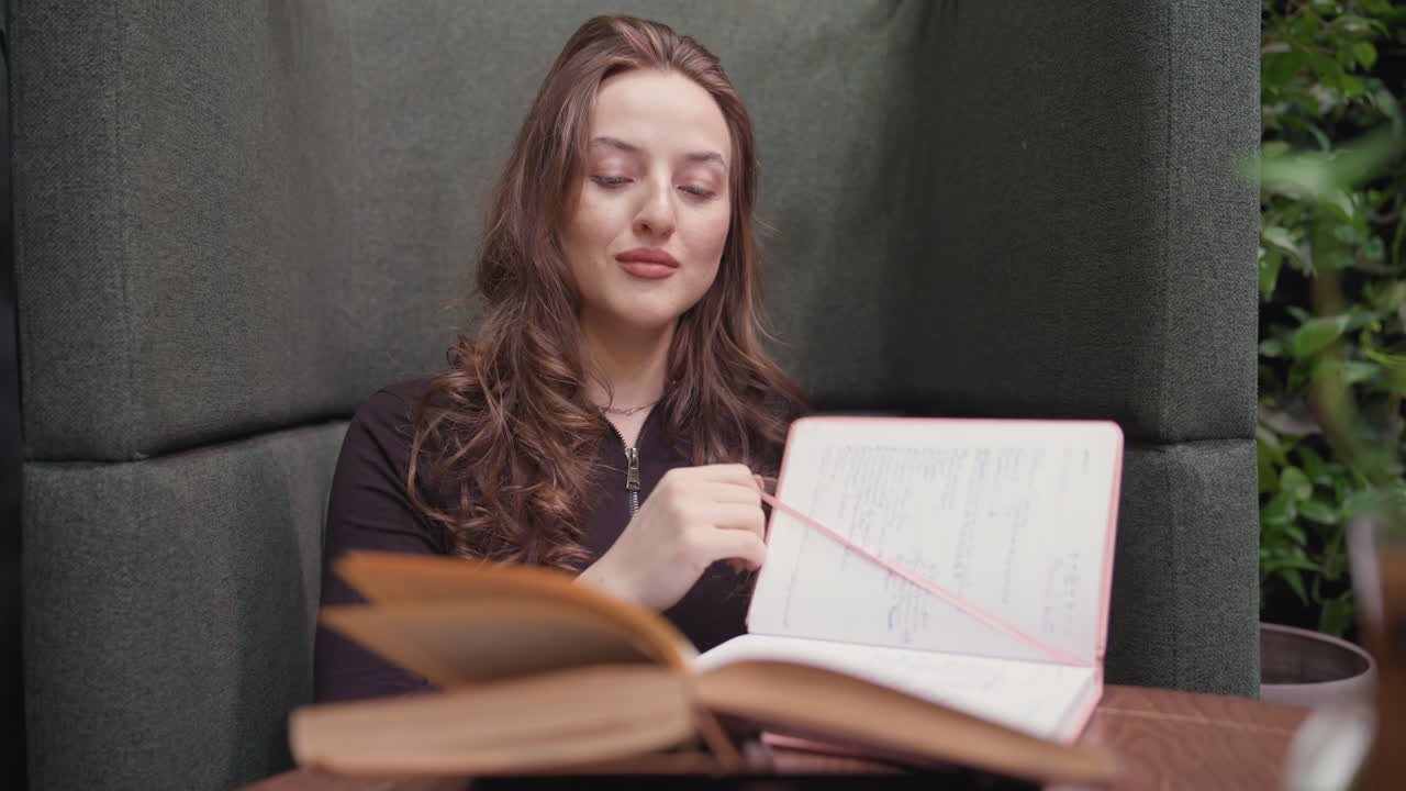 Lady adjusts sleeve while seated on cushion in quiet indoor setting, preparing to open book on wooden table surrounded by soft green decoration and relaxed workspace vibe