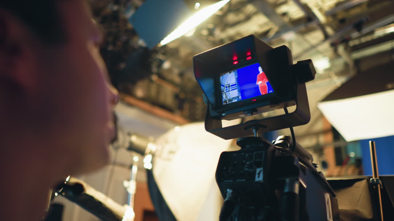 Cameraman monitors professional camera screen while filming female presenter in red dress on blue screen stage during studio shoot, surrounded by lighting equipment, teleprompter, and production gear in active setup