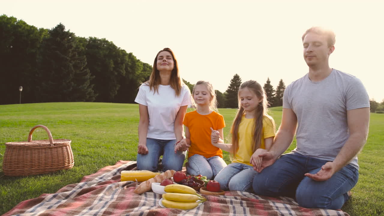 felices padres e hijas con los ojos cerrados arrodillados en el prado, tomados de la mano y meditando durante un picnic en el parque 1