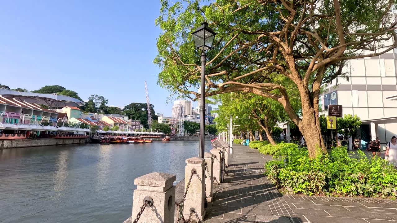 Daytime camera pan along Clarke Quay riverfront walkway, lush greenery, waterfront buildings, clear sky