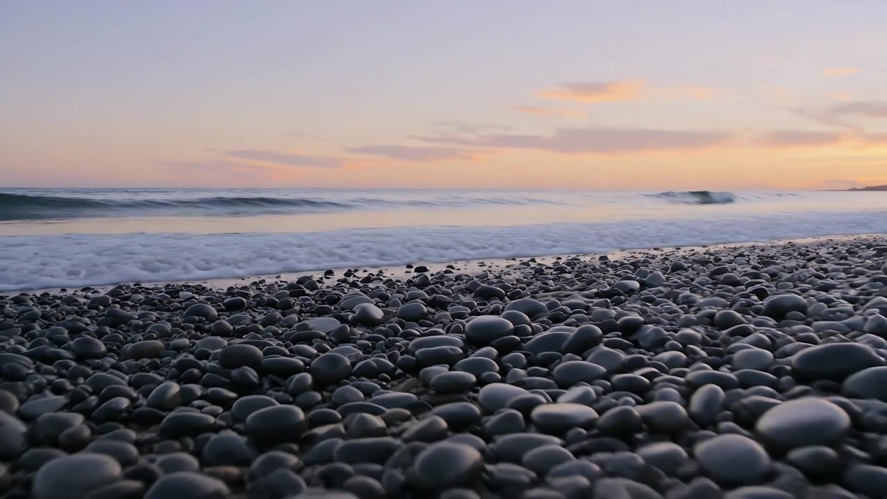 Onda che si avvicina da destra a una costa di ciottoli all'alba, pietre nere bagnate che luccicano