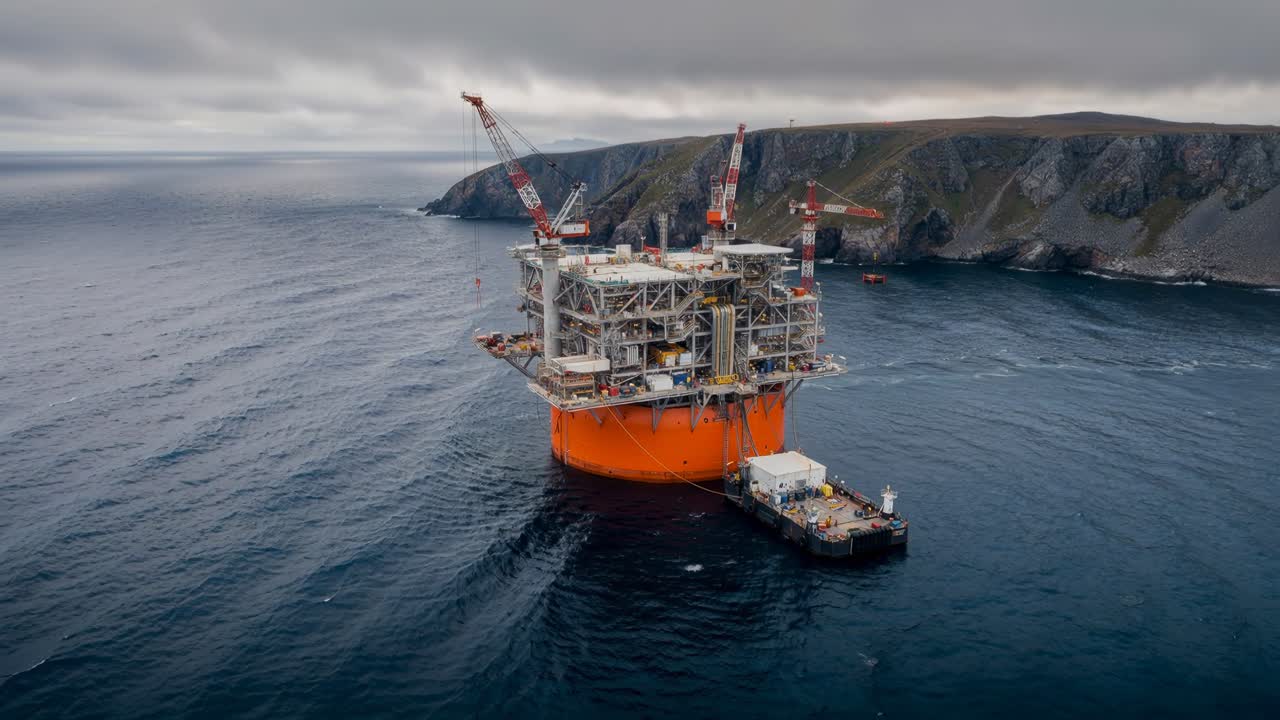 Luftfoto af en offshore olieplatform med servicepram i roligt hav nær barske kystklipper under overskyet himmel.