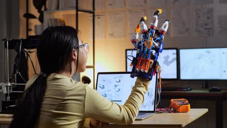 back view of teen asian girl with 3d printing checking a cyborg hand while designing it on a laptop at home