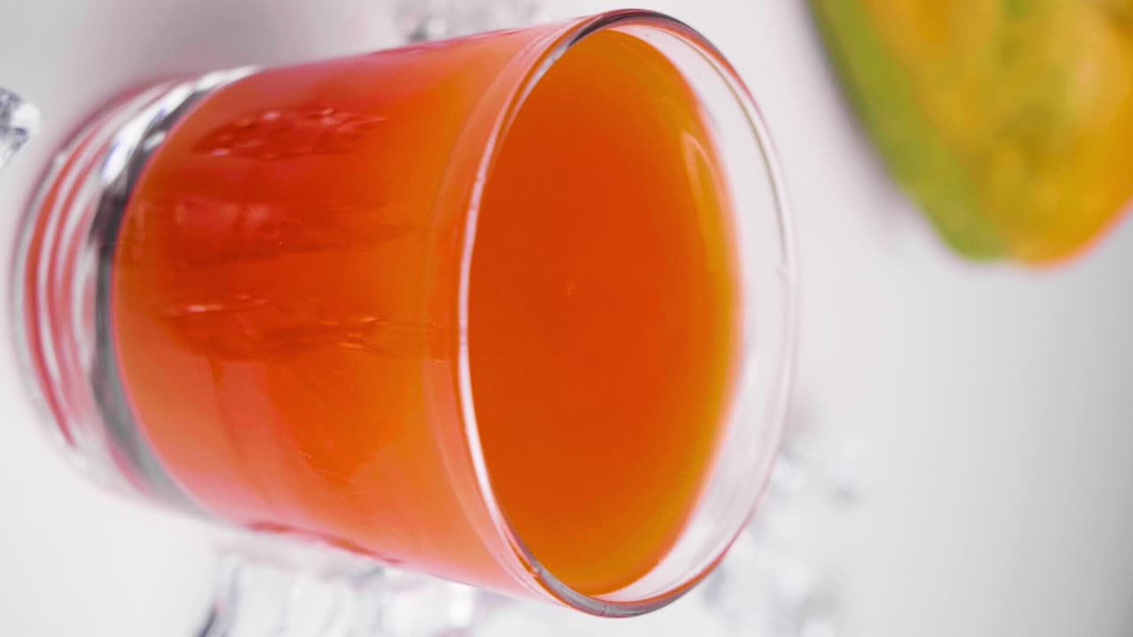 Ripresa verticale al rallentatore di succo di papaya in un bicchiere con uno schizzo causato da una goccia che cade e una papaya sullo sfondo