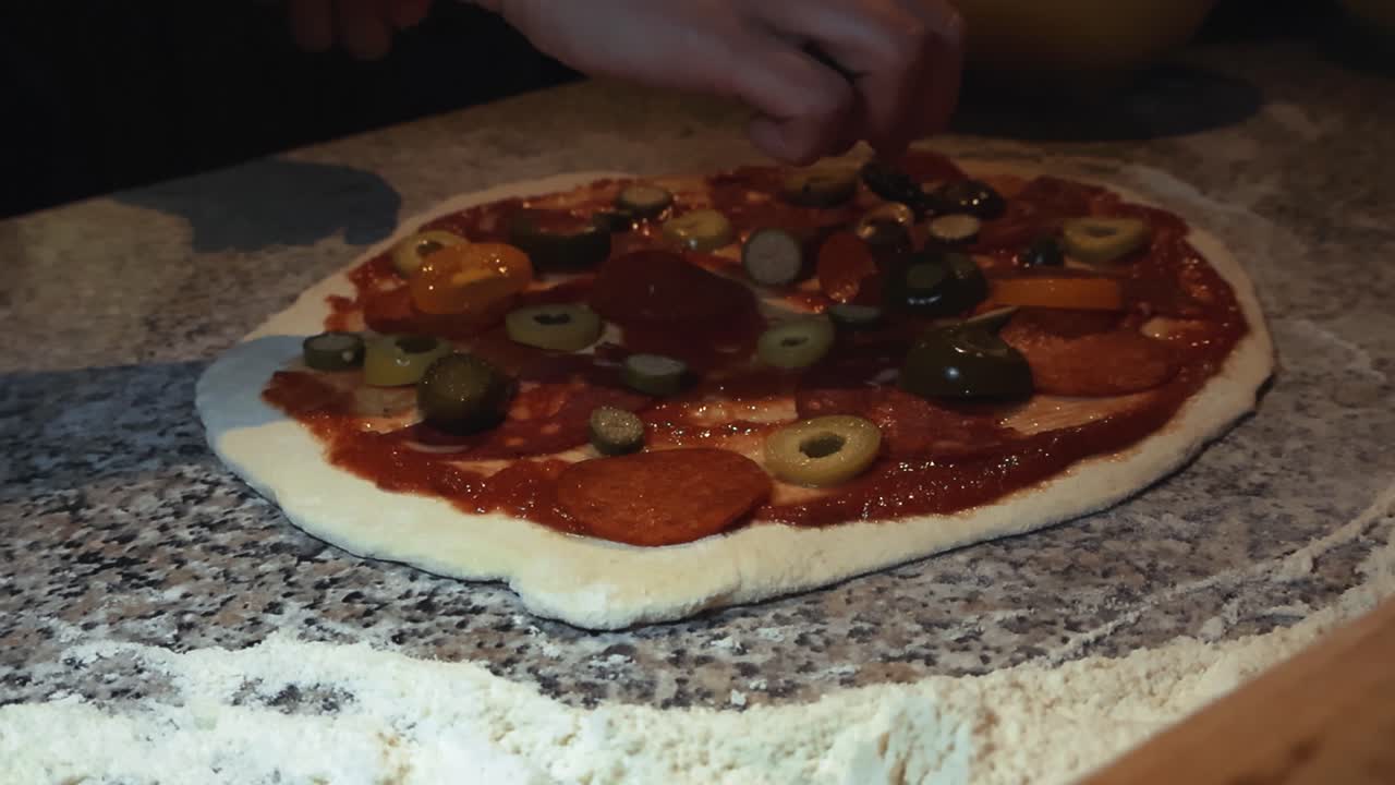 Close up slow motion video of a diverse white chef hand placing chorizo and other toppings on a freshly rolled and made pizza dough that has tomato sauce and olives on it. White flour, home kitchen